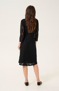 Robe en dentelle noire avec des manches trois quarts, motif floral, longueur au genou, tissu texturé et ourlet fini. Portée avec des chaussures noires.