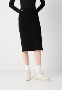 Personne portant une jupe noire côtelée longueur genou avec des chaussettes blanches et des baskets blanches, debout devant un fond clair uni.