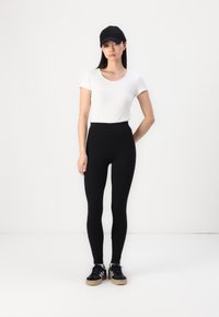 Zwarte high-waisted leggings van rekbare stof, gecombineerd met een aansluitend wit T-shirt en een zwarte baseballcap, gedragen met zwarte sneakers.