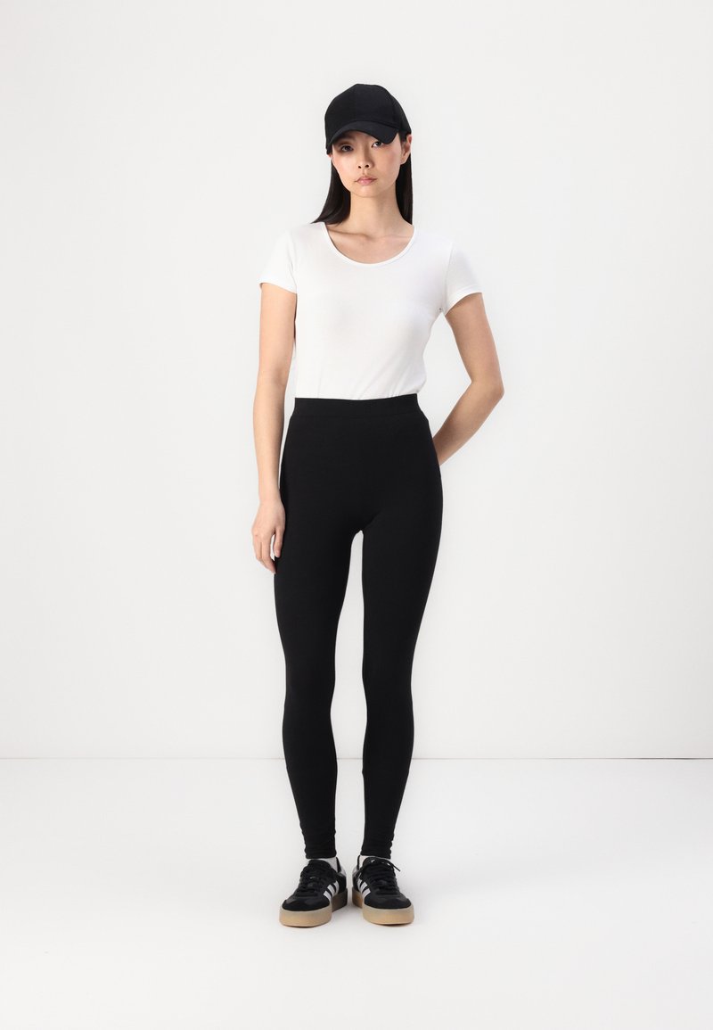 Zwarte high-waisted leggings van rekbare stof, gecombineerd met een aansluitend wit T-shirt en een zwarte baseballcap, gedragen met zwarte sneakers.