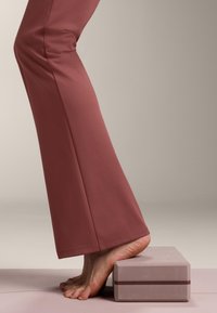 Persona con pantaloni color ruggine in equilibrio sulle punte dei piedi su un blocco yoga color malva su un tappetino da esercizio viola chiaro.