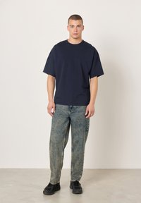 Ohlapna mornarsko modra majica iz bombaža. Par svetlo modrih posivelih kavbojk z ravnim krojem. Črni supergi dopolnjujejo videz.