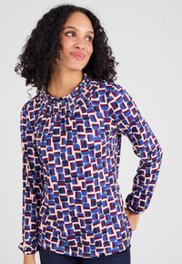 Blusa a maniche lunghe con scollo arricciato, realizzata in tessuto leggero con un motivo geometrico multicolore in blu, rosso e crema.