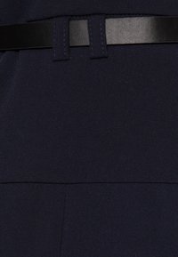 Tissu bleu marine foncé à texture lisse, avec une ceinture noire à la taille. Des coutures visibles ajoutent du détail à la structure du vêtement.