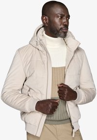 Beige polstret jakke med hætte, lynlås og ribbede manchetter. Laget over en hvid og olivengrøn tekstureret striktrøje.