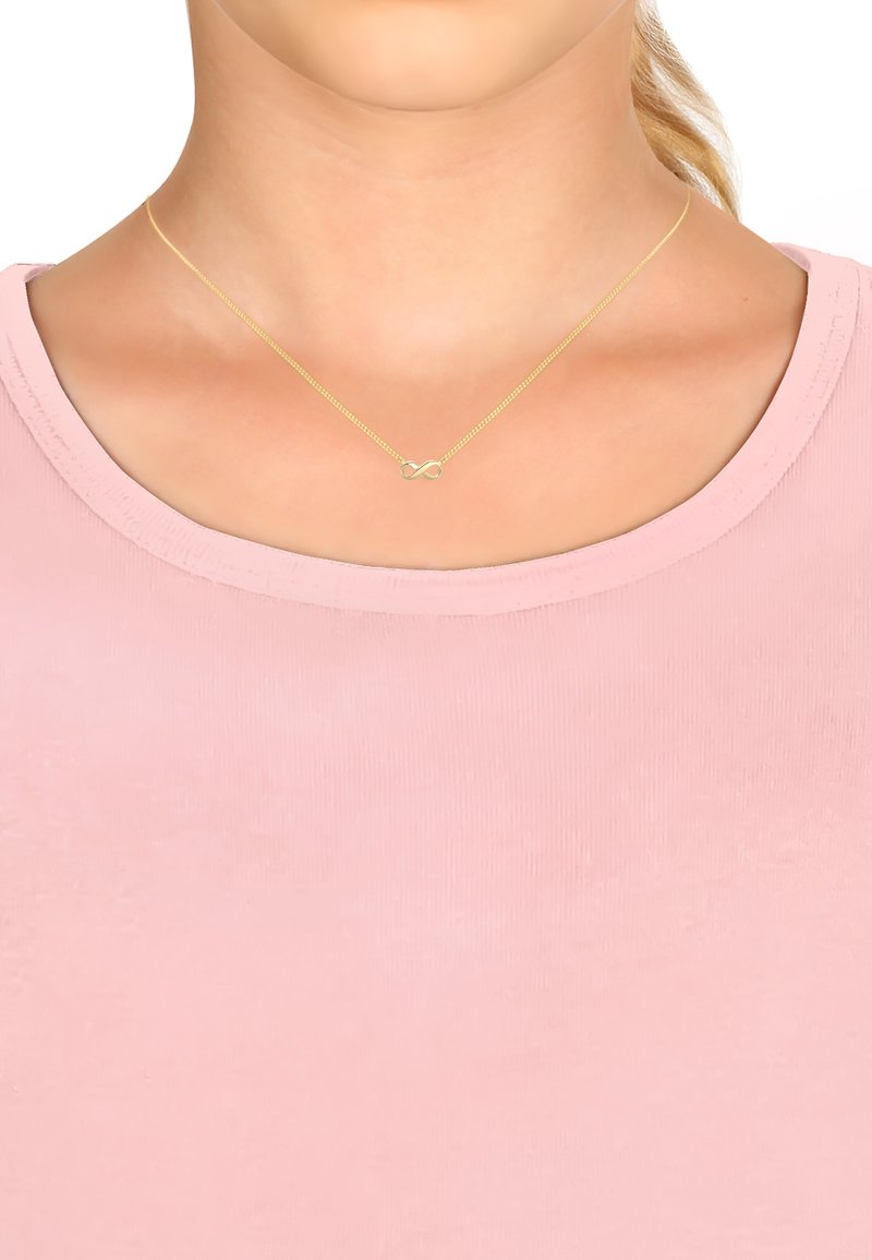 Guldhalskæde med et uendelighedssymbol vedhæng, der har en tynd kæde og glat overflade, båret over en lys pink top.