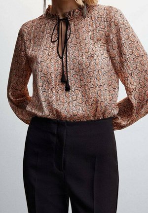 Vrouw draagt een blouse met lange mouwen en bloemenprint, met een zwarte strik bij de hals, ingestopt in zwart, hooggetailleerd, op maat gemaakt broek.