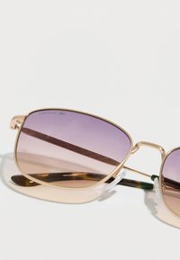 Ochelari de soare cu rame metalice aurii, lentile degrade mov, accente texturate maro pe brațe și detalii verzi pe tije. Logo Lacoste vizibil.