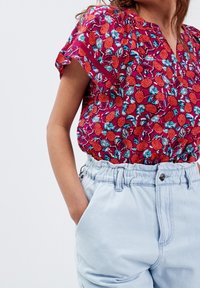 Donna con una mano nella tasca di jeans chiari a vita alta, indossa una maglietta a maniche corte fucsia con un motivo floreale rosso e turchese.