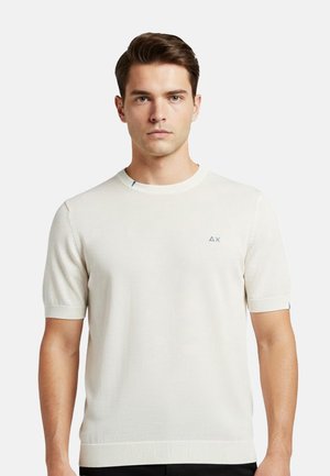 Giovane uomo che indossa una maglietta a maniche corte in maglia color avorio chiaro con un piccolo logo sul petto sinistro, in piedi davanti a uno sfondo uniforme.