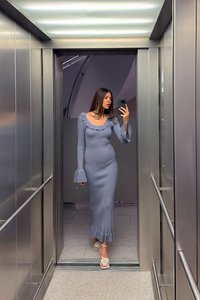 Langes, figurbetontes Kleid aus weich gestricktem Stoff in sanftem Blau mit rüschigem Ausschnitt und weit ausgestellten Ärmeln. Mit einem schwingenden Saum und leichten, hellfarbenen Schuhen ausgestattet.