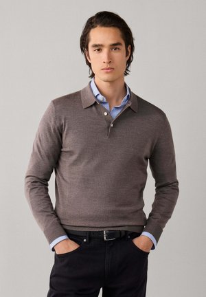 Joven con cabello oscuro que lleva un polo marrón de manga larga sobre una camisa azul a rayas, pantalones negros y cinturón negro, con las manos en los bolsillos.
