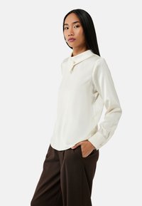 Camicia bianca con collo e una sola chiusura a bottone, maniche lunghe e una texture liscia, abbinata a pantaloni marroni.