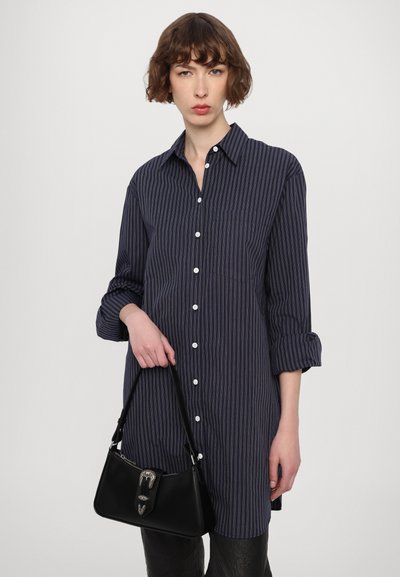 Camisa a rayas azul marino con cierre de botones, mangas enrolladas y un bolsillo frontal. Sosteniendo un pequeño bolso de cuero negro con un cierre plateado.