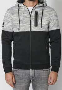 Felpa con zip nera e grigia, con una sezione superiore testurizzata, una sezione inferiore di colore solido a contrasto, tasche laterali e una tasca con zip sul petto.