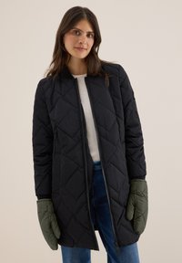 Černý prošívaný dlouhý kabát se zapínáním na zip, kulatým výstřihem a pružnými manžetami. Zelené rukavice doplňují outfit. Model má na sobě modré džíny.