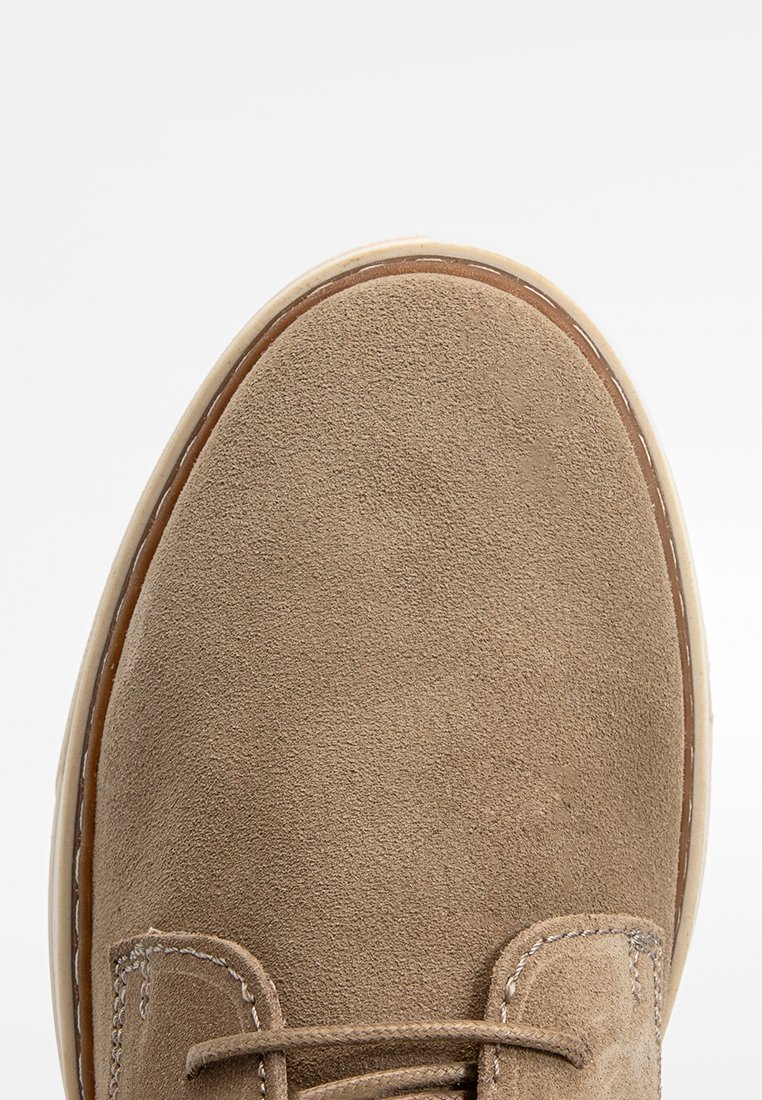 Beige Wildlederschuh mit runder Zehenpartie, sichtbarem Nähen, passenden Schnürsenkeln und einer leichten Gummisohle. Glatte Textur ohne auffällige Verzierungen.