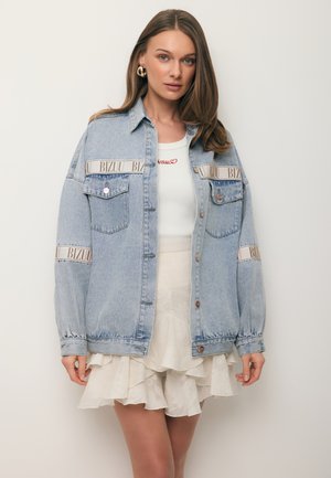 Vrouw draagt een oversized lichtblauwe spijkerjasje met "BIZUU" tekstbanden, een wit topje en een gelaagde witte ruchesrok, staand tegen een effen achtergrond.