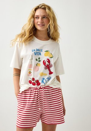 Ung kvinde iført en hvid T-shirt med teksten "La Dolce Vita" og madillustrationer samt rødhvide stribede shorts, smilende og kigger til siden.