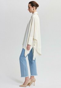Poncho blanc texturé avec col et liens sur les côtés, associé à un jean bleu clair avec des ourlets effilochés et des escarpins beiges à bouts pointus.