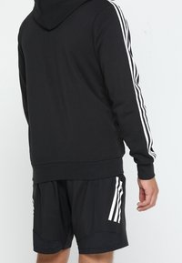 Sudadera negra con capucha, que presenta tres franjas blancas a lo largo de cada manga. Combinada con pantalones cortos negros, también decorados con franjas blancas.