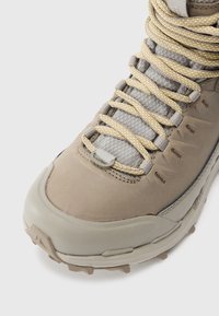 Botte de randonnée couleur tan avec dessus texturé, lacets jaunes et gris, et semelle en caoutchouc. Comprend un bout renforcé et un col rembourré pour le soutien.