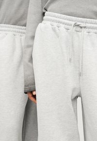 Ανοιχτό γκρι φόρμα από μαλακό ύφασμα, με ελαστική μέση και κορδόνι. Μερικώς ορατό υφασμάτινο γκρι μακρυμάνικο μπλουζάκι με υφή.