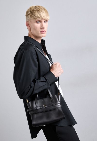 Une personne blonde vêtue d'une chemise et d'un pantalon noirs tient un sac à main en cuir noir avec une bandoulière contre un fond gris clair.