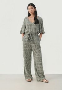 Ženska stoji z rokami v žepih, oblečena v vzorčast siv kombinezon s širokimi hlačnicami in kratkimi rokavi, v kombinaciji z belimi sandali.