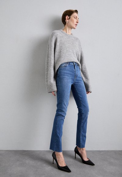 Gray knitted sweater with wide sleeves, paired with high-waisted blue jeans featuring frayed hems and black pointed-toe heels.