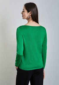 Pull vert à manches longues avec un large décolleté, texture lisse, coupe ajustée et ourlet côtelé. Porté avec un pantalon noir et des boucles d'oreilles en argent.