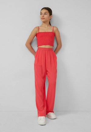 Giovane donna in piedi contro un muro grigio, indossa un top corto rosso, pantaloni rossi a vita alta e sneakers bianche, mani nelle tasche, guarda verso l'alto.