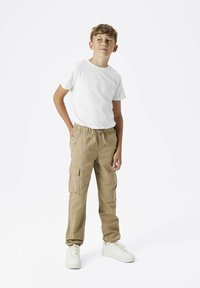 Pantalones cargo beige con cintura elástica y bolsillos laterales, combinados con una camiseta blanca lisa y zapatillas blancas, sobre un fondo neutro.