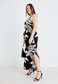 Robe maxi noire avec imprimé feuilles tropicales en crème et bordeaux. Elle présente une taille à nouer, une fente sur le côté et des bretelles spaghetti réglables. Associée à des talons noirs.