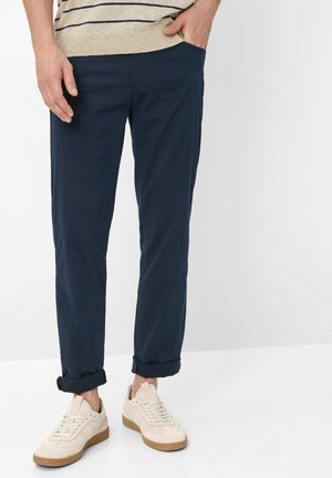 Mężczyzna ubrany w granatowe spodnie ze ściągaczami, beżowy sweter w paski oraz kremowe tenisówki z gumowymi podeszwami, stojący na tle białego tła.