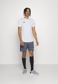 Baltas sporta T-krekls ar īsām piedurknēm un melnu logo, apvienots ar pelēkajiem šortiem un melnām augstās zābakām līdz ceļiem. Pelēki sporta apavi ar sietiņa dizainu.
