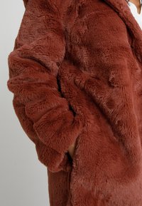 Ruda spalvos dirbtinė kailio striukė, turinti minkštą, pūkuotą tekstūrą, pasižyminti didelėmis kišenėmis ir laisvu siluetu. Dizainas apima plačias rankoves.