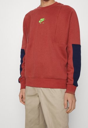 Sudadera roja con mangas azul marino, que presenta un logo de Nike verde en el pecho. Está hecha de un material suave con un corte relajado y acentos texturizados.