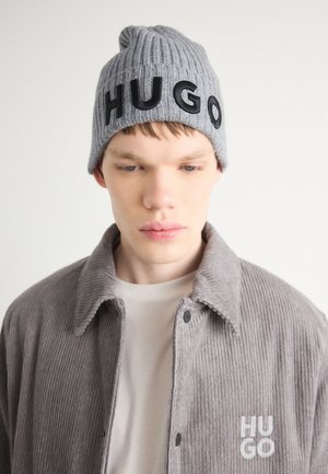 Gestrickte graue Mütze mit "HUGO" in schwarzen Buchstaben. Das Modell trägt eine graue Cord-Jacke mit einem dezent gestickten "HUGO"-Logo auf der Brust.
