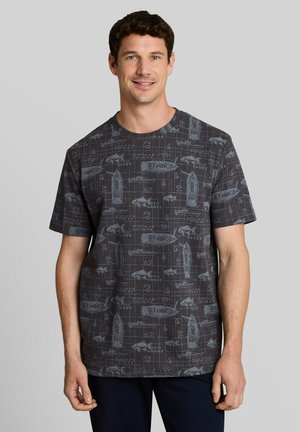 Gelijkende man met kort donker haar die een donkergrijs T-shirt draagt met nautische boot- en vissenblauwdrukpatronen, staand tegen een effen achtergrond.
