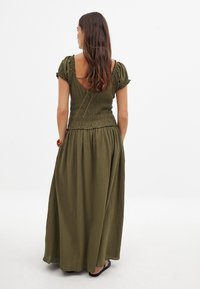 Olivgrünes Maxikleid mit kurzen Puffärmeln und gesmokter Taille. Es verfügt über einen offenen Rücken und einen fließenden Rock, der bis zu den Knöcheln reicht.