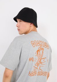 Gray cotton T-shirt featuring an orange graphic on the back with text "DICKIES LAND AHOY" and an illustrated character. Black ribbed bucket hat.