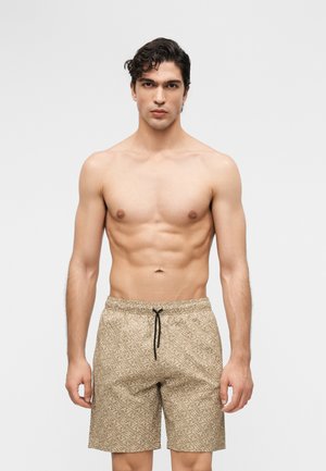 Homme torse nu aux cheveux courts et bouclés, portant un short de bain beige à motifs avec un cordon noir, debout devant un fond blanc uni.