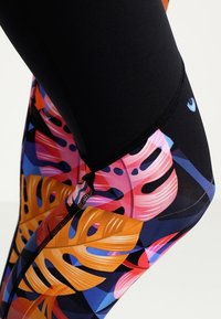 Legginsy z jaskrawym, tropikalnym wzorem liści w odcieniach pomarańczowego, różowego i niebieskiego na czarnym tle z widocznymi detalami szwów.