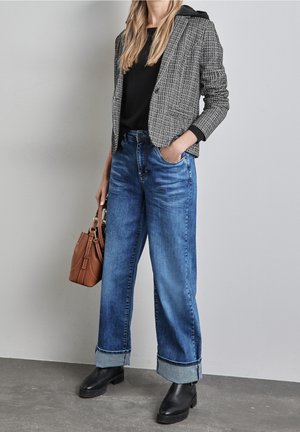 Vrouw draagt een zwarte top, grijze geruite blazer met capuchon, omgeslagen wijde blauwe jeans, zwarte laarzen, houdt een bruine handtas vast tegen een effen muur.