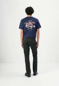 Camiseta gráfica azul marino con la impresión "PARA EL VIAJE DE LA VIDA EST. 1947", combinada con jeans oscuros y zapatos negros, vista desde la parte de atrás.