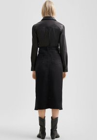 Blonde vrouw in een zwart leren shirt met lange mouwen, zwarte hoge taille midi rok en zwarte enkellaarzen, staand tegen een effen achtergrond.