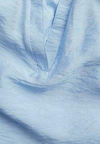 Helles blaues Gewebe mit einer weichen, strukturierten Oberfläche, das einen tiefen V-Ausschnitt und subtile Falten aufweist, was auf einen lässigen Stil hinweist.