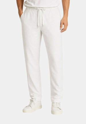 Uomo che indossa pantaloni bianchi con coulisse e sneakers bianche, in piedi con le mani nelle tasche su uno sfondo semplice.