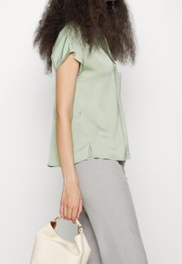 Grön satin kortärmad topp med v-ringning och sidofickor, kombinerad med ljusgrå vida byxor och en krämfärgad handväska.